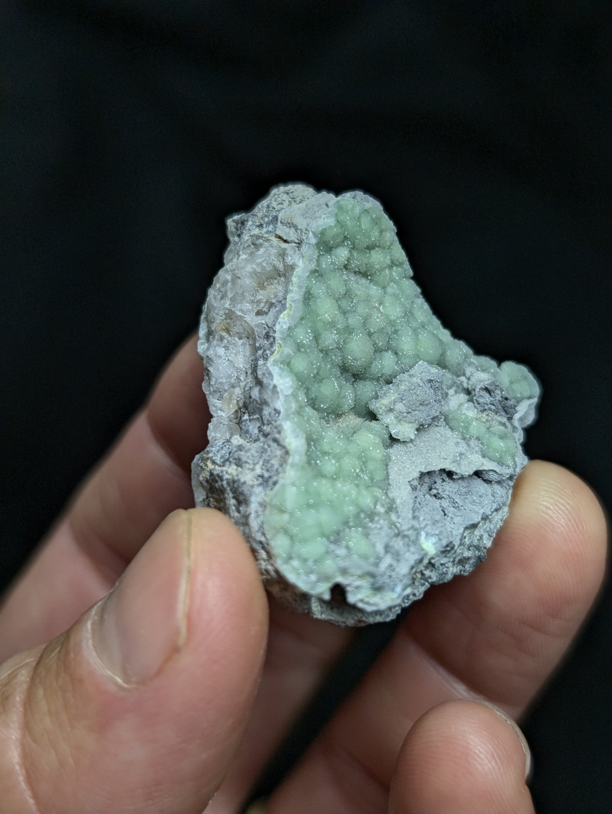 A hand holds a rough, irregularly shaped mineral specimen featuring clusters of pale green crystals growing from a grayish matrix.