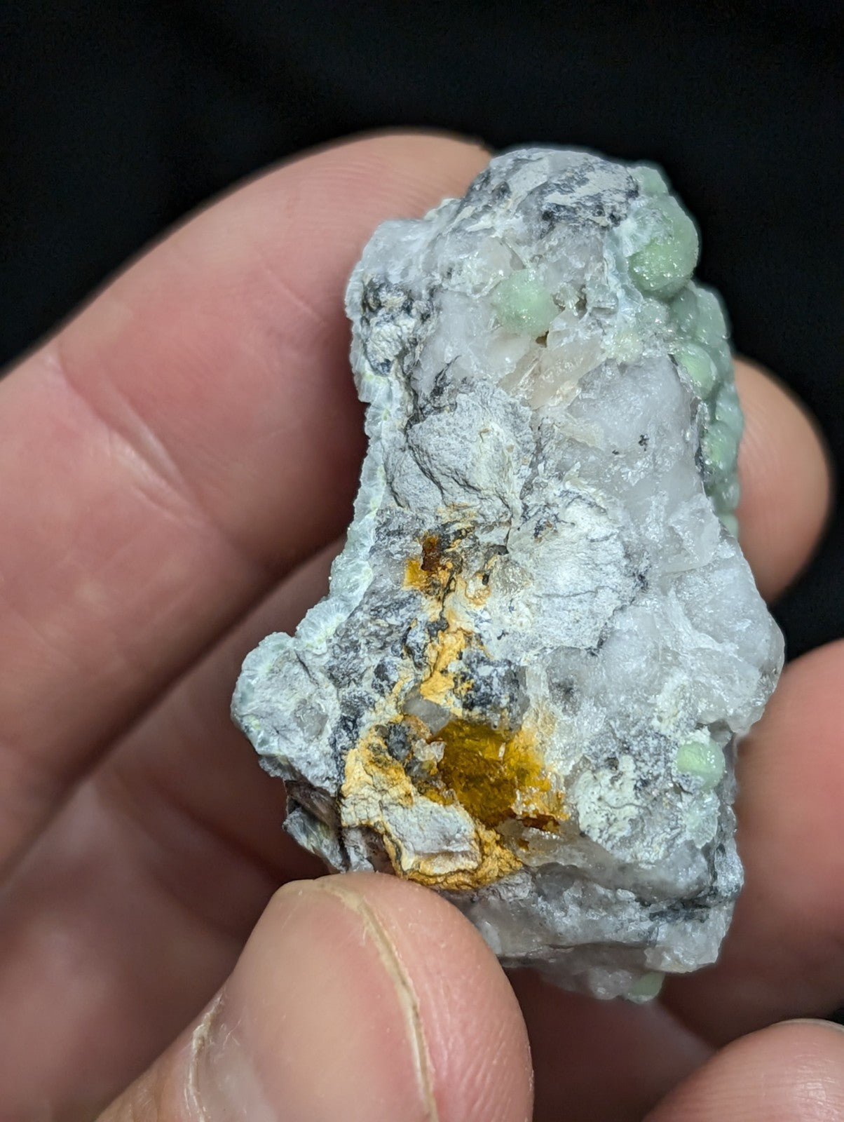 A rough, irregularly shaped mineral specimen with a pale gray matrix featuring patches of translucent green crystals and areas of bright yellow-orange mineralization.