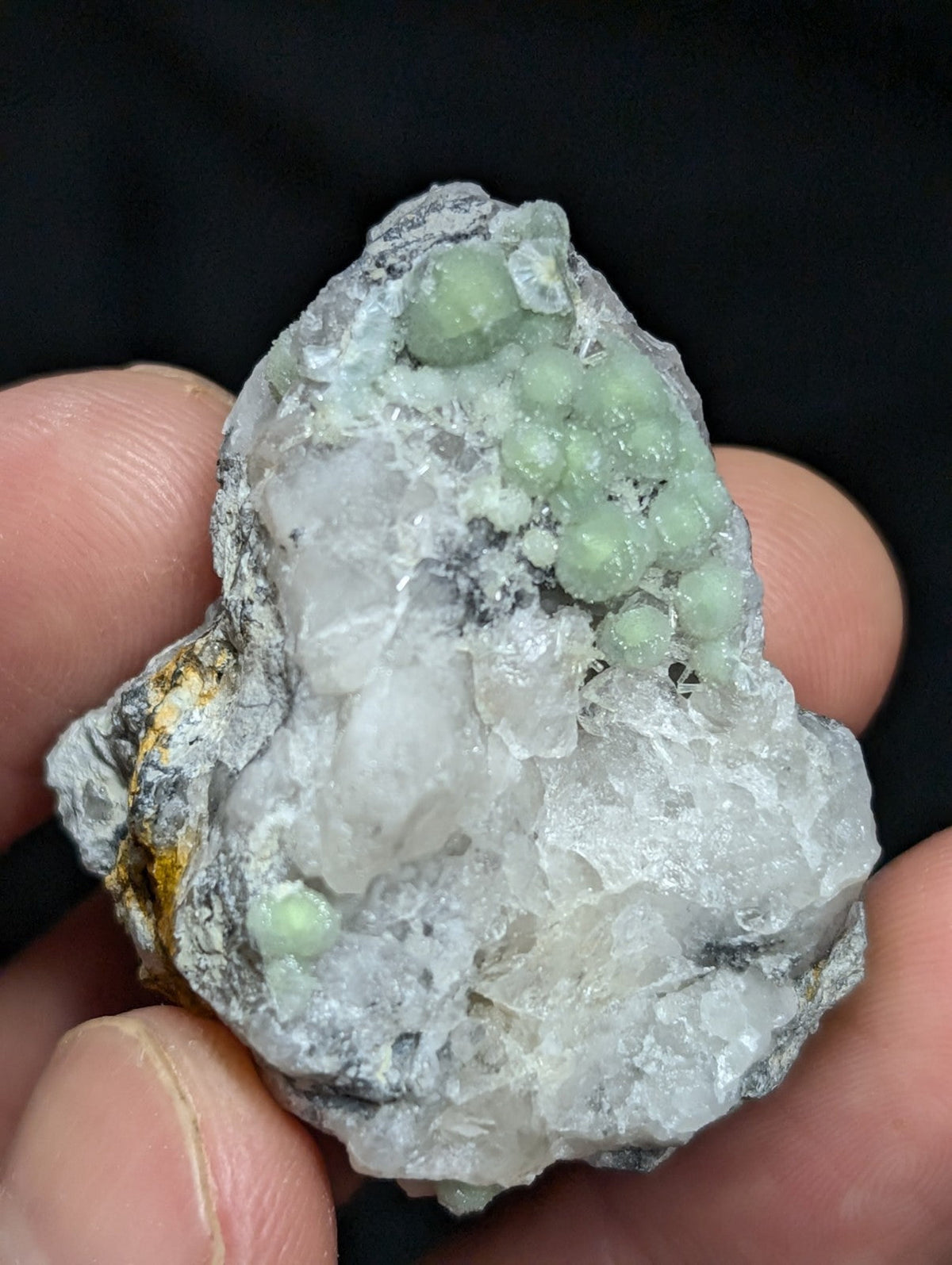 A hand holds a rough, irregularly shaped mineral specimen featuring translucent white crystals with clusters of pale green spherical formations.