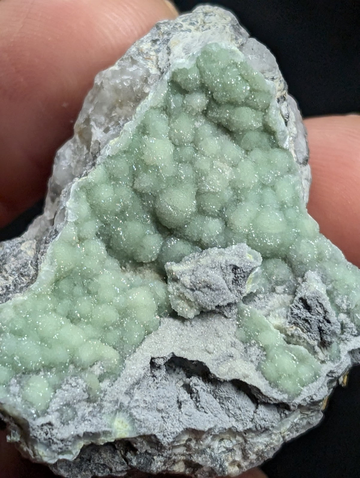 A hand holds a rough, gray rock specimen featuring clusters of translucent, light green crystals with a sparkling, glittery surface.