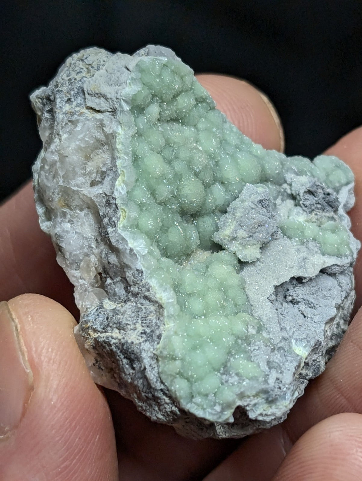 A hand holds a geode with translucent, light green crystalline formations nestled within a rough gray rock matrix.