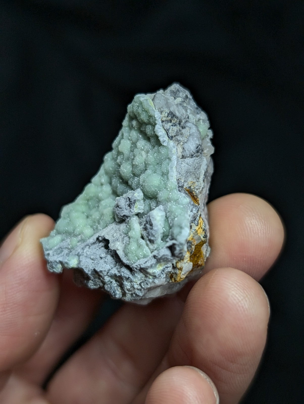 A textured, light green mineral specimen with a crystalline surface and patches of gray matrix, accented by small golden flecks.