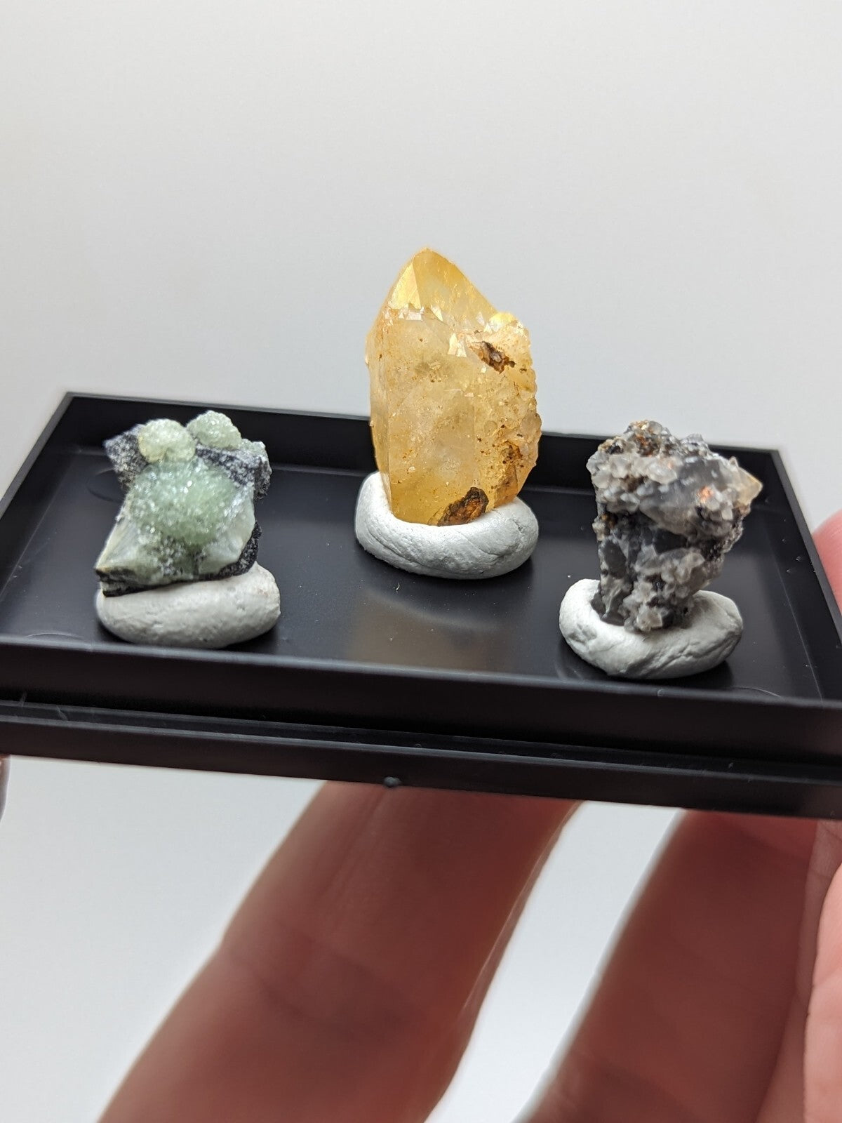 A black tray holding three distinct mineral specimens, each mounted on a white base.