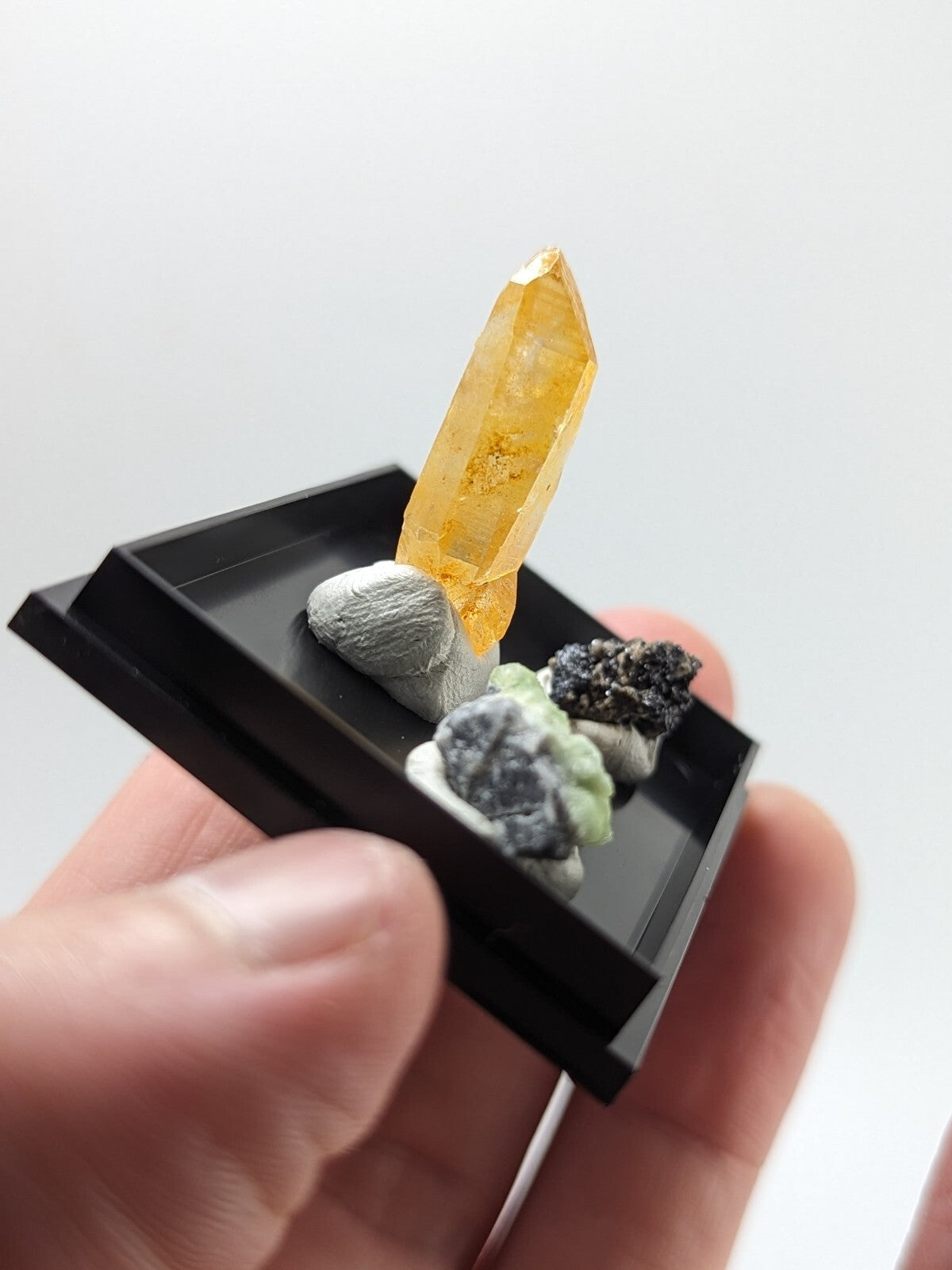 A vibrant yellow-orange crystal with a translucent, faceted structure stands prominently in a sleek black display tray alongside smaller mineral specimens.