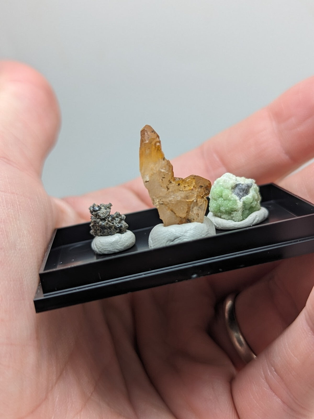 Black rectangular display tray holding three miniature mineral specimens on white ceramic bases.