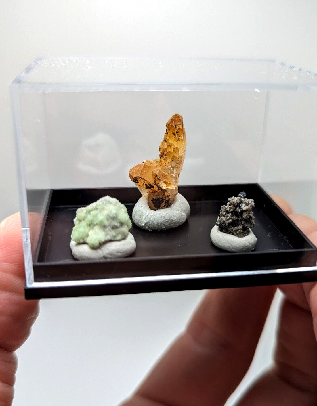 透明展示盒内陈列着三件矿物标本，中央为橙黄色晶簇、左侧是浅绿色团块状矿石、右侧则是深灰色颗粒状矿石。