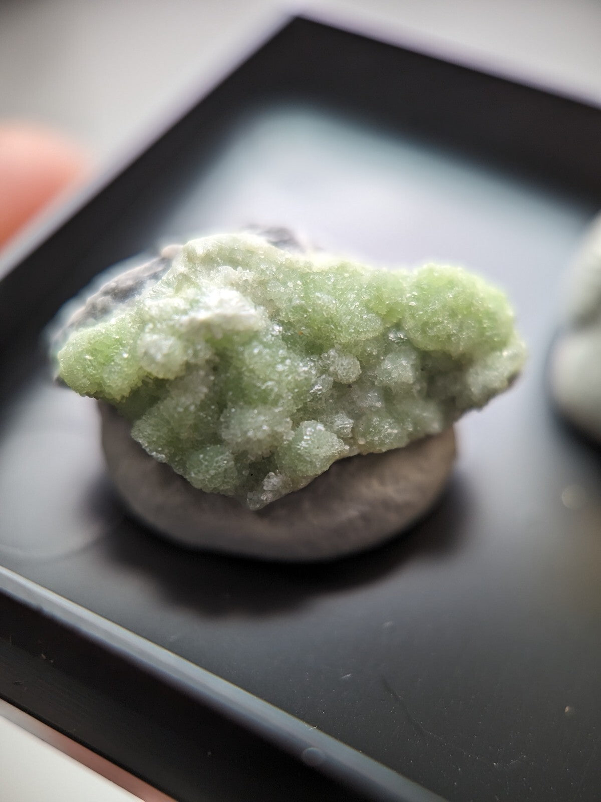 A vibrant green, crystalline mineral specimen with a textured, bumpy surface rests atop a smooth gray stone.