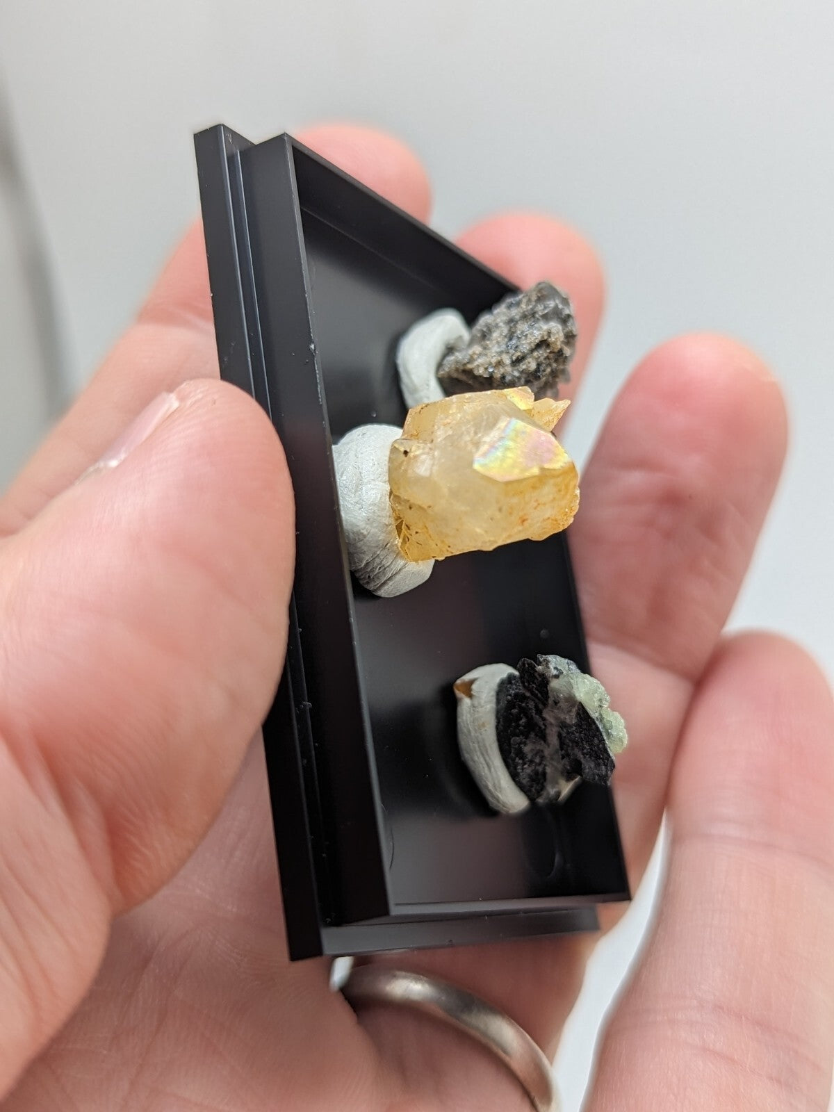 Black rectangular mineral display case holding a collection of small, naturally occurring rocks and crystals.