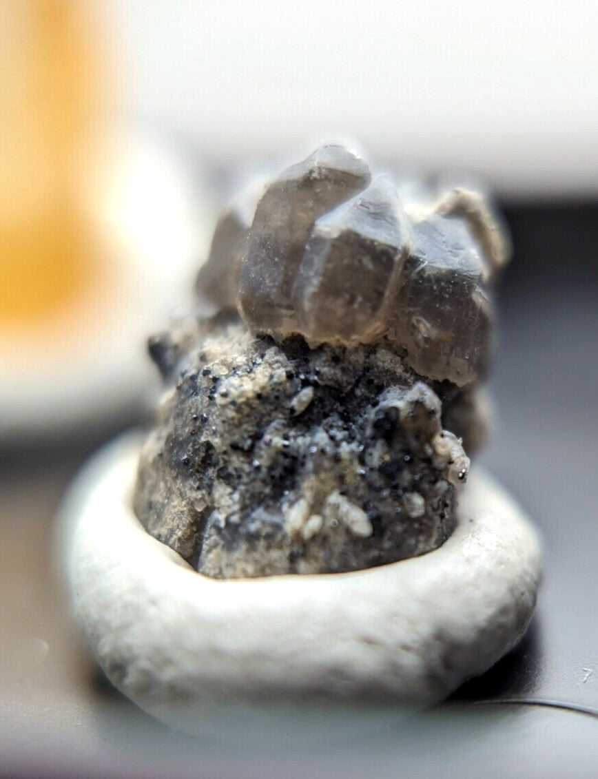 A cluster of translucent brownish-gray quartz crystals with a rough, earthy base rests in a small white ceramic dish.