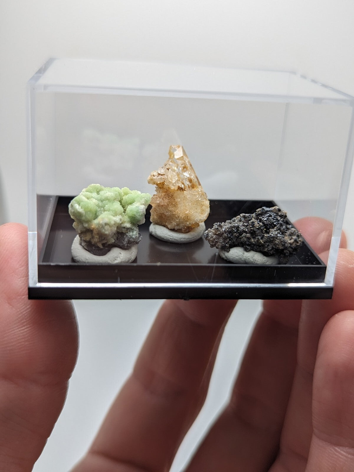 A clear acrylic display case holding three distinct mineral specimens on a black base.