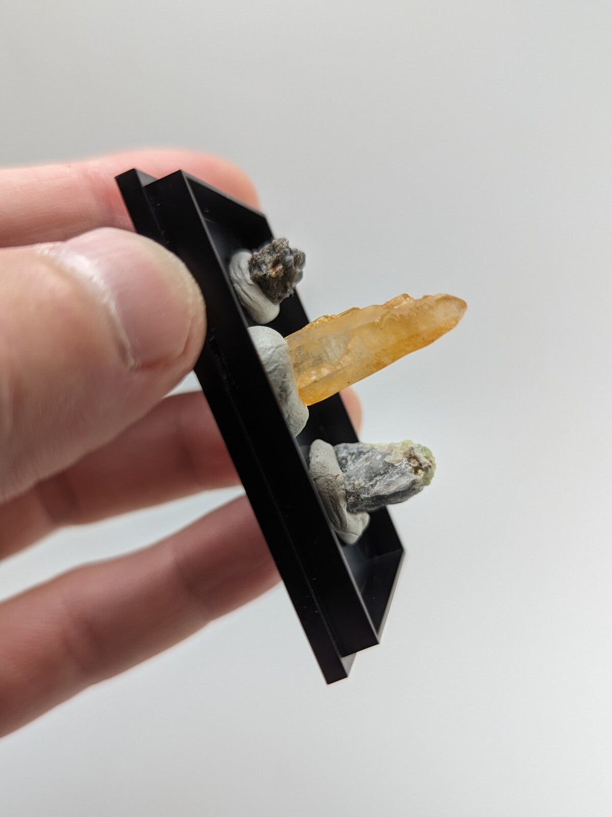 A black rectangular display case holding three mineral specimens, including a translucent yellow crystal and two grayish rock samples.