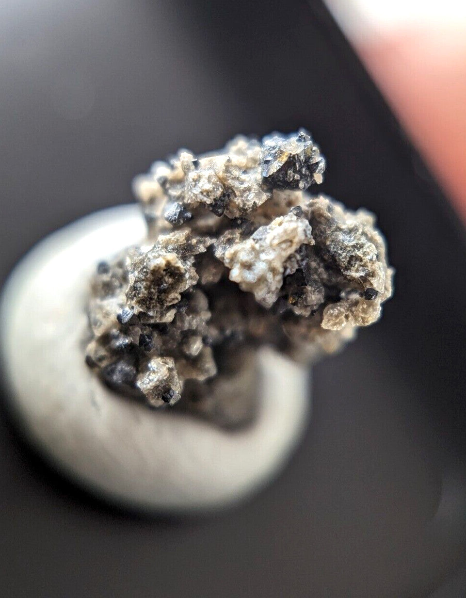 A rough, clustered mineral specimen with a mix of gray and black tones sits atop a smooth white circular base.