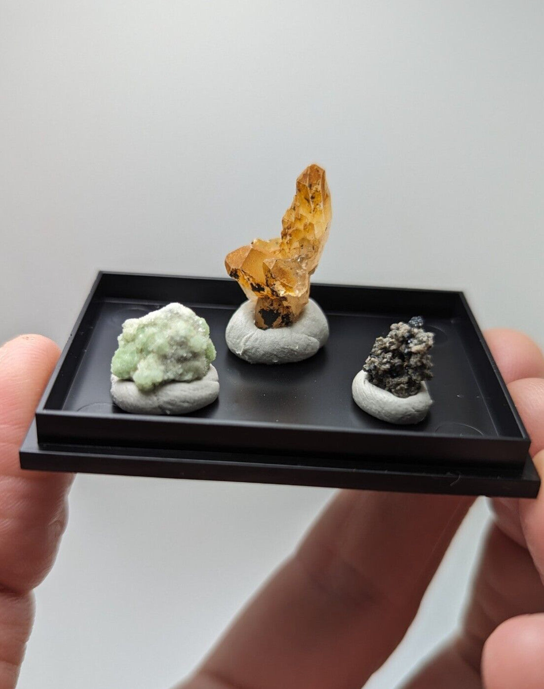 Black rectangular display tray holding three mineral specimens on individual white stone bases.