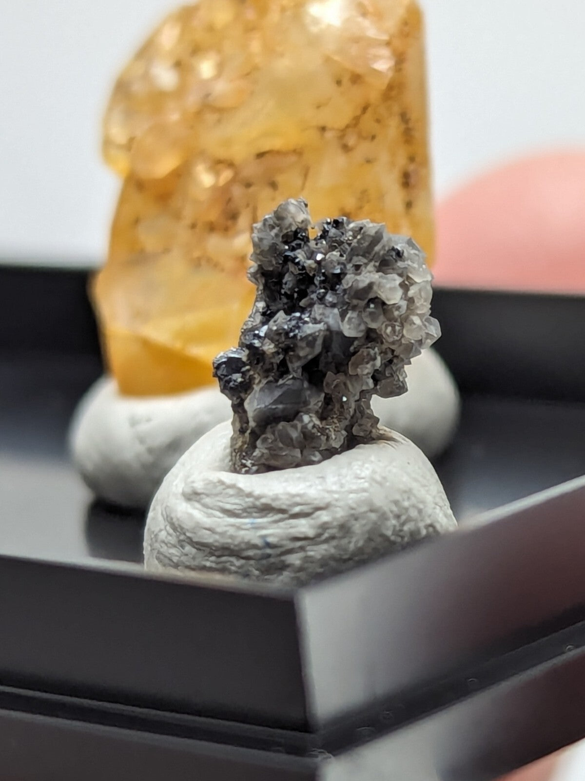 A gray and black mineral specimen with crystalline formations sits atop a textured white base.