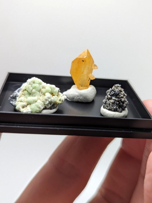 A miniature black display tray holding three distinct mineral specimens: a pale green, textured rock; a vibrant yellow-orange crystal with sharp facets resting on a white base; and a dark gray, mossy-looking mineral also set in a white pedestal.