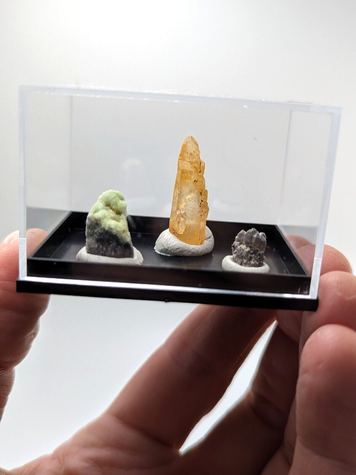 A clear acrylic display case holding three distinct mineral specimens: a green and gray rock, an amber-colored crystal with visible inclusions on a white base, and another small dark gray rocky specimen.
