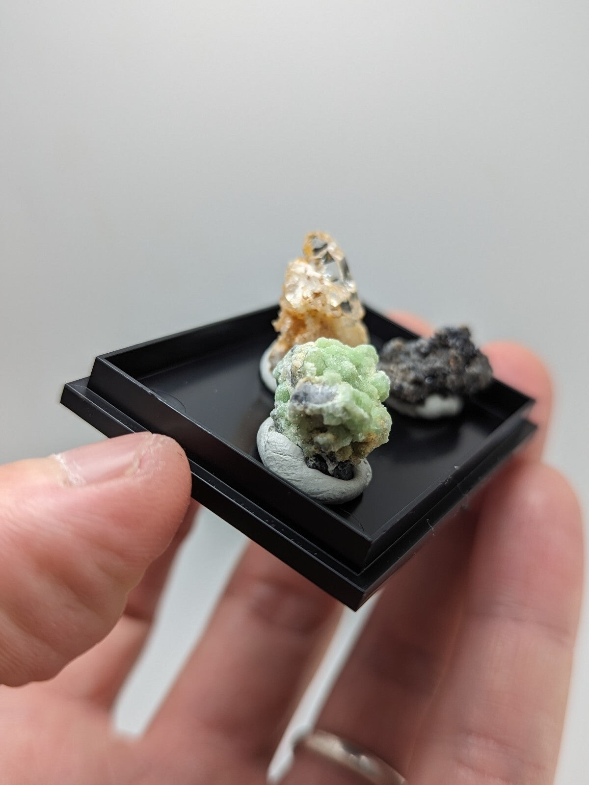 Black rectangular display tray holding three miniature mineral specimens with varied colors and textures.