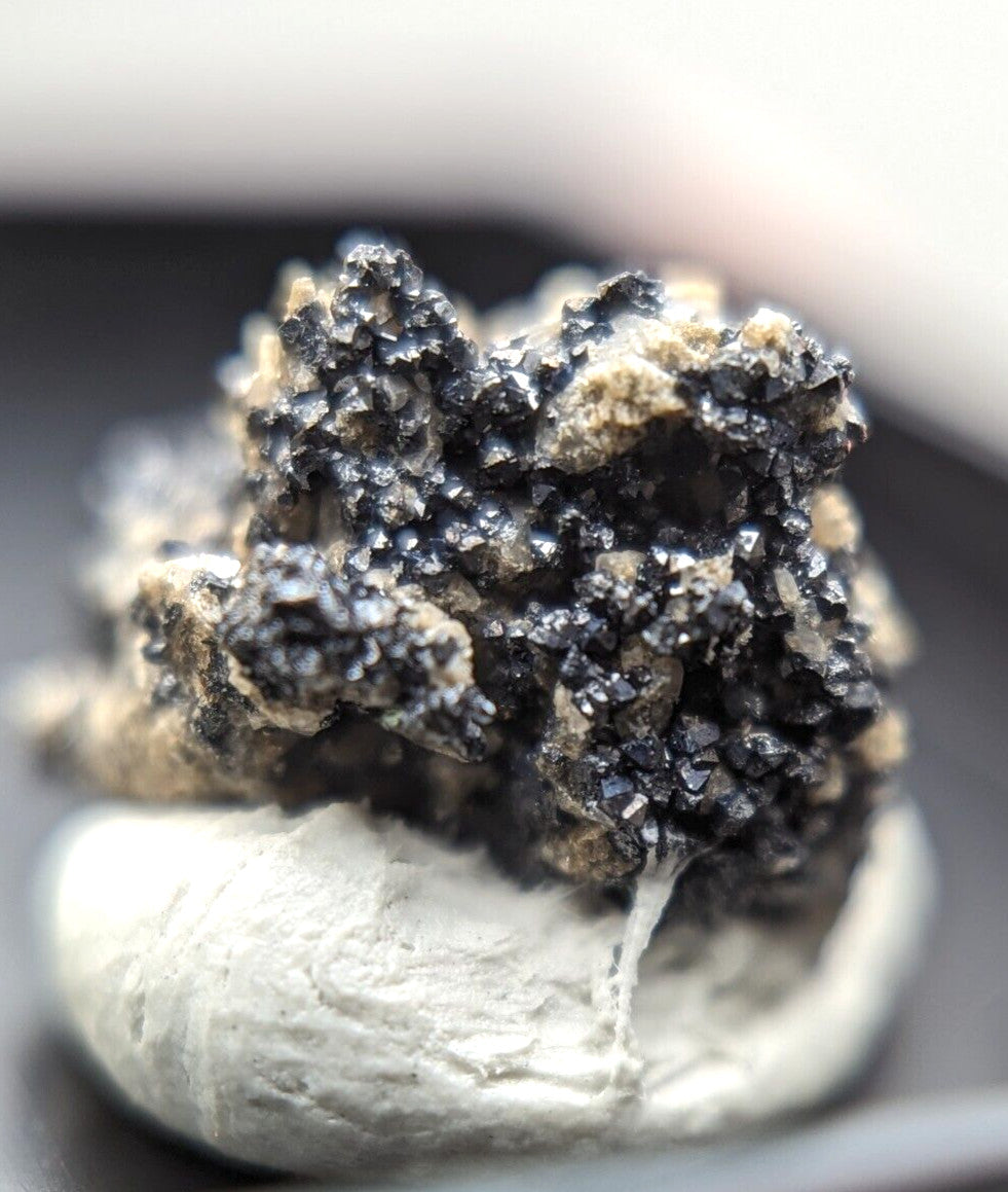 A cluster of dark, crystalline minerals with sharp facets and a metallic sheen rests atop a smooth, light gray rock base.