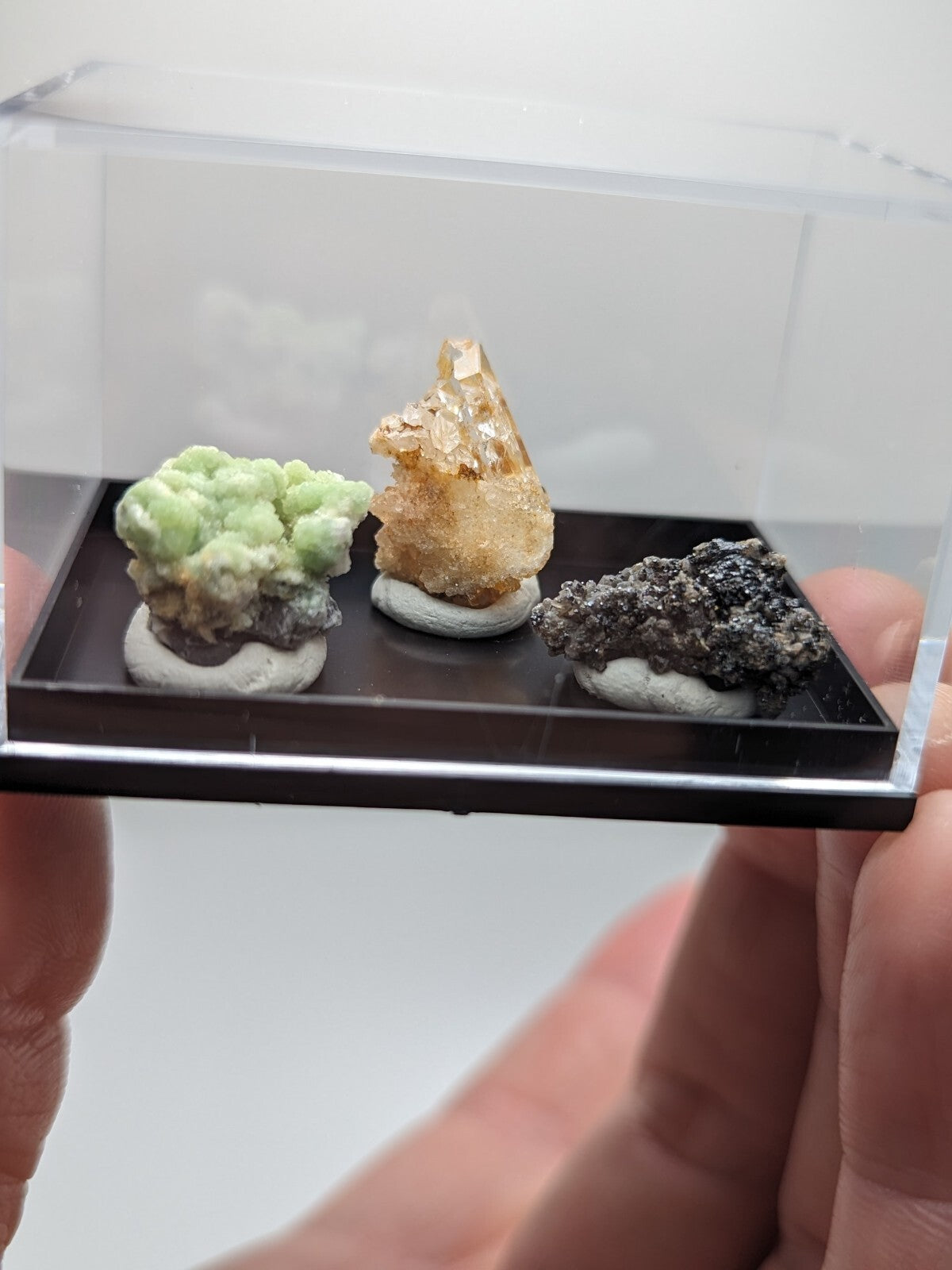 A clear acrylic display case holding three distinct mineral specimens, each mounted on a white circular base.