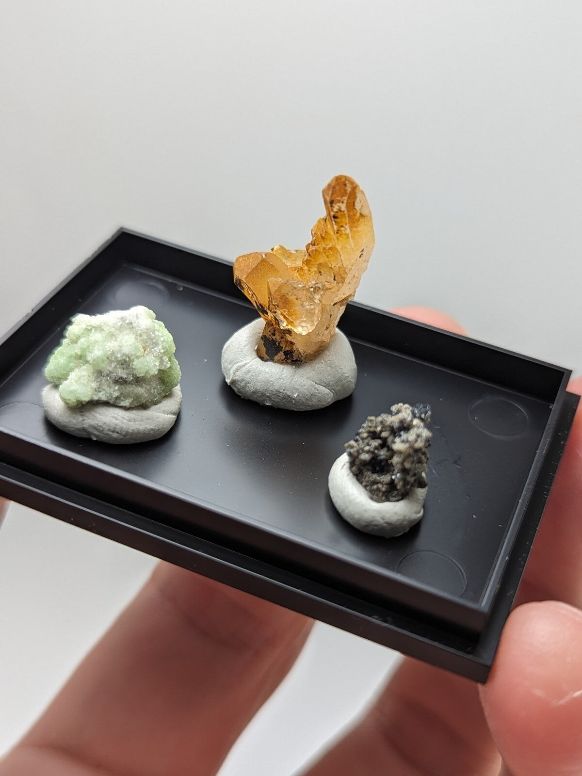 A black rectangular tray holding three miniature mineral specimens, each mounted on a white circular base.