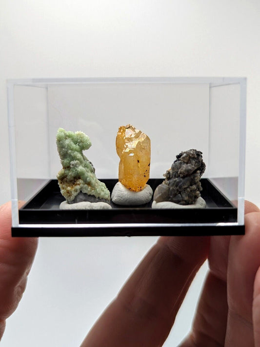 A clear acrylic display case holding three distinct mineral specimens: a light green crystalline formation, a translucent golden-yellow crystal on a white base, and a dark grayish-black rock with speckled texture.