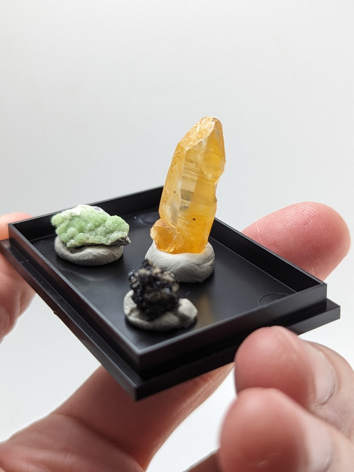 A miniature black display tray holding three small mineral specimens: a green crystalline formation, an amber-yellow translucent crystal with visible internal striations on a gray base, and a dark metallic-looking specimen also resting on a gray base.