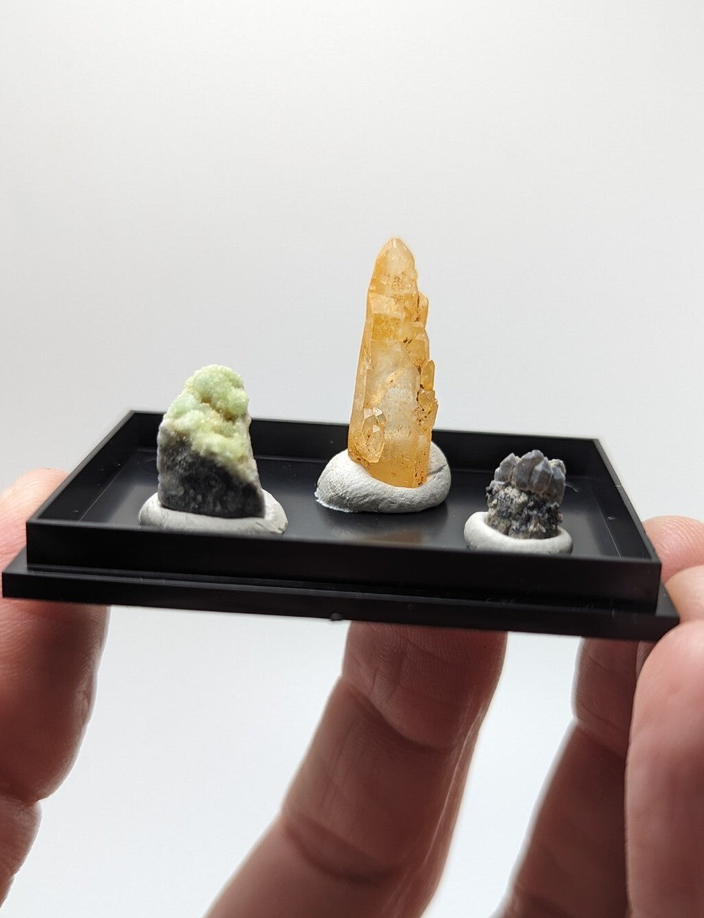 Black rectangular display tray holding three mineral specimens on white circular bases.
