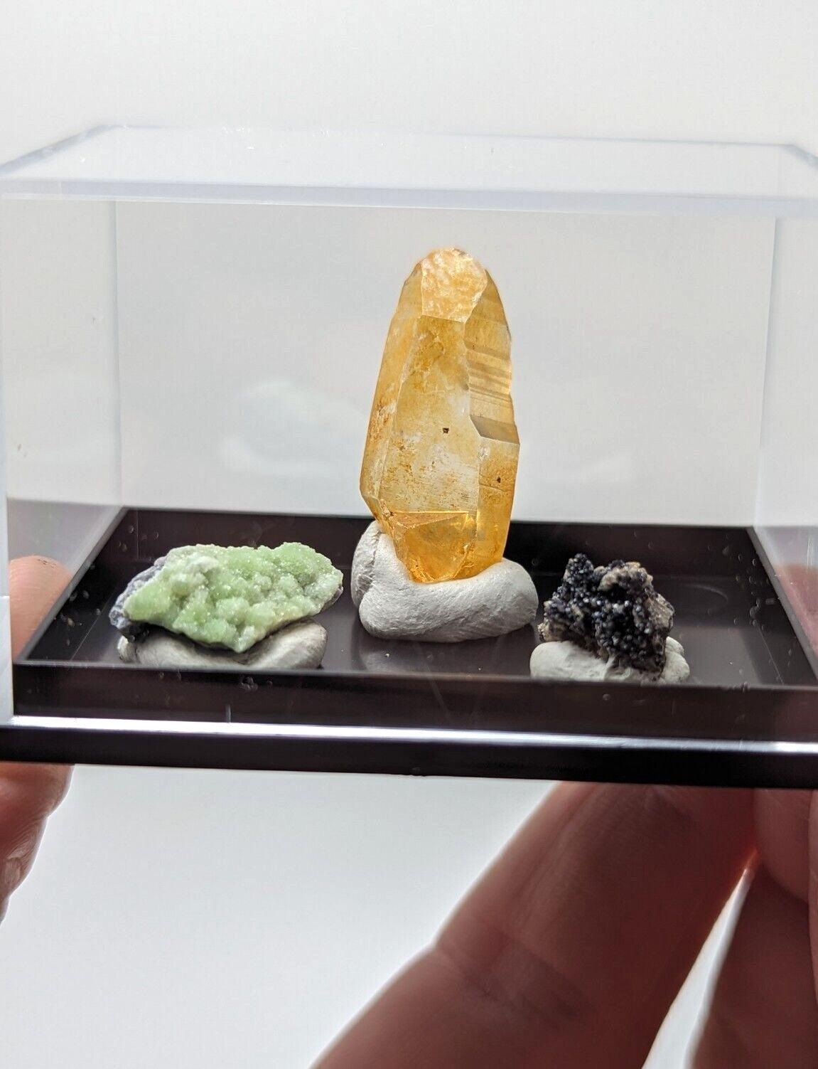 A translucent golden-yellow crystal specimen with a faceted, pointed top and visible internal inclusions is displayed upright on a small white rock base inside a clear acrylic display case.