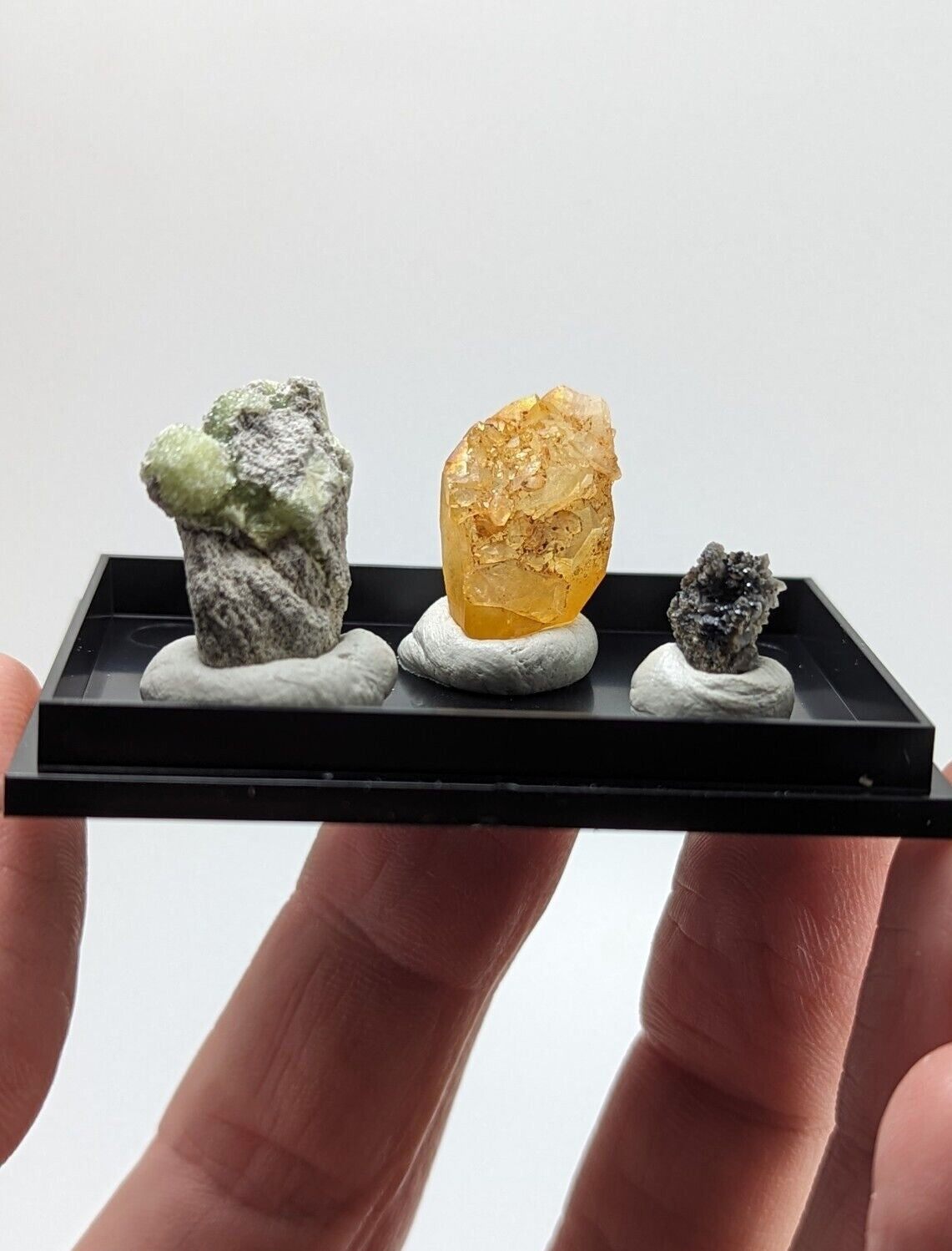 A miniature black display tray holding three distinct mineral specimens, each mounted on a white base.