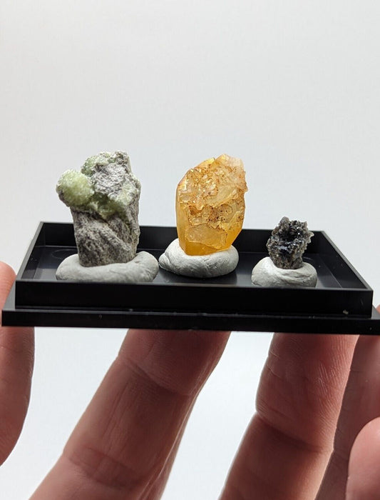 A miniature black display tray holding three distinct mineral specimens, each mounted on a white base.