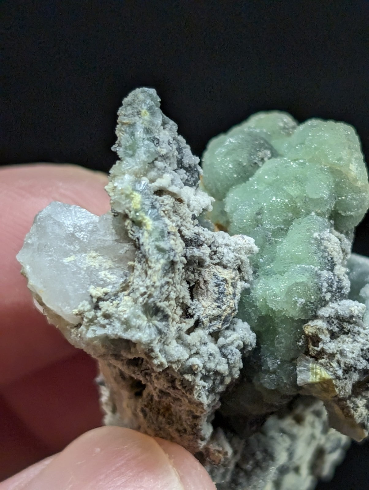 A hand holds a mineral specimen featuring translucent green crystals growing from a rough, grayish matrix.