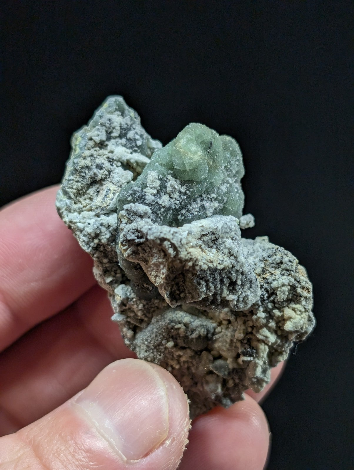 A rough, irregularly shaped green mineral specimen with a crystalline texture and speckled grayish matrix.