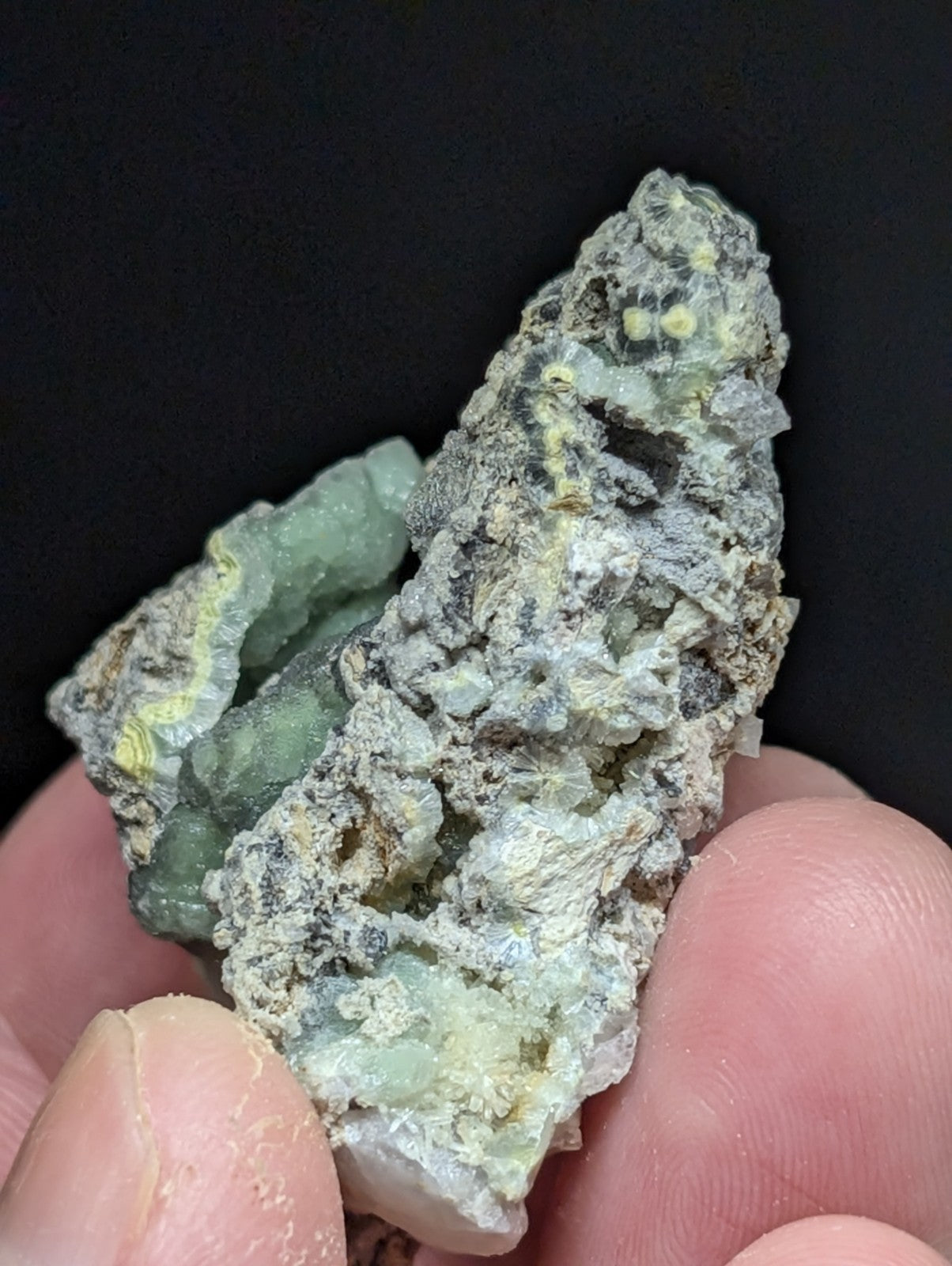 A rough, irregularly shaped mineral specimen with a mix of green and gray hues, featuring translucent crystalline formations embedded in a darker matrix.
