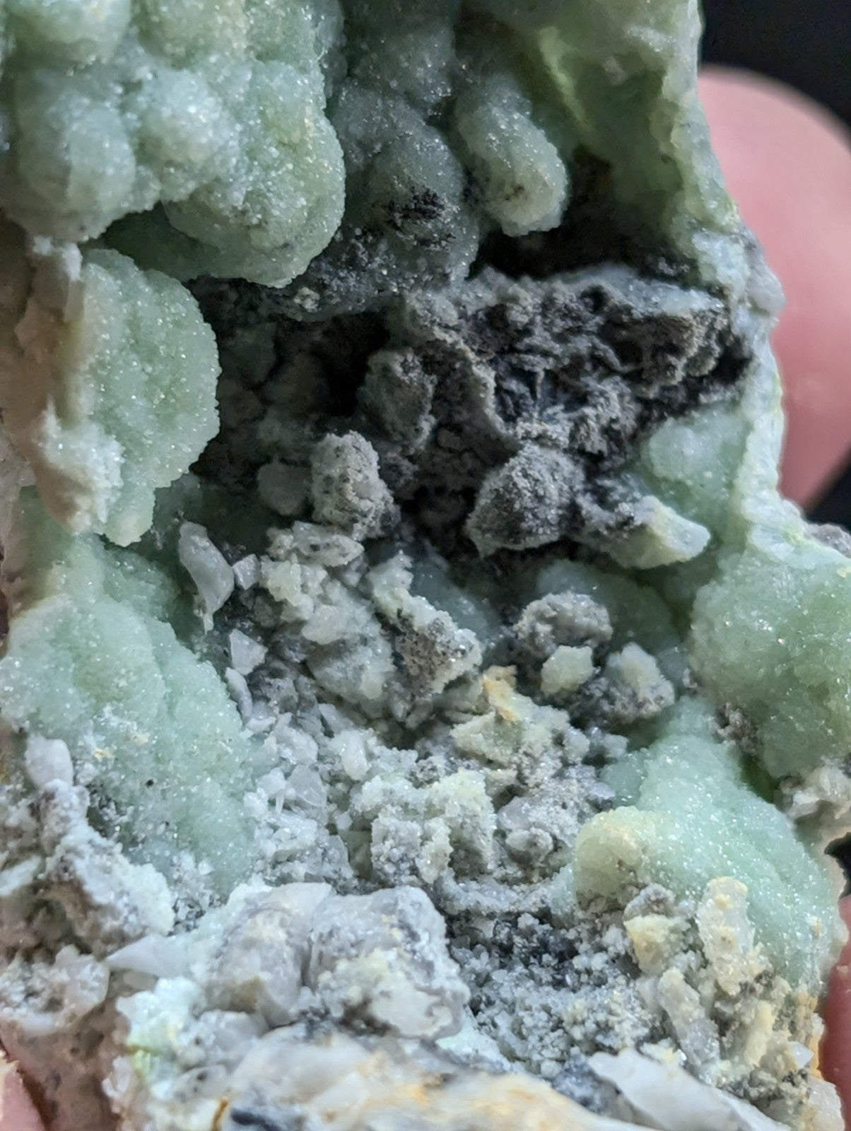 A pale green, crystalline mineral specimen with a rough, textured surface and embedded darker grayish-black inclusions.