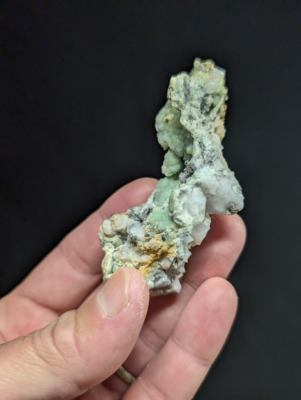 A hand holds a rough, irregularly shaped mineral specimen featuring patches of pale green and white with streaks of yellowish-orange.