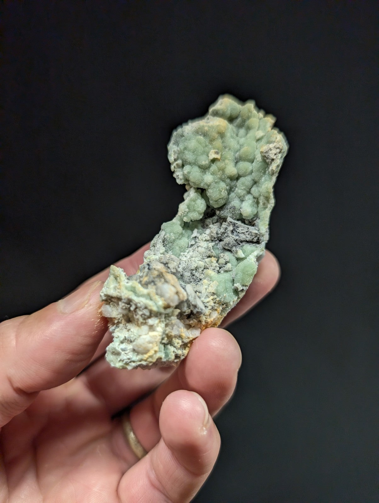 A hand holds a rough, irregularly shaped mineral specimen with a mottled green and gray surface featuring clustered crystalline formations.