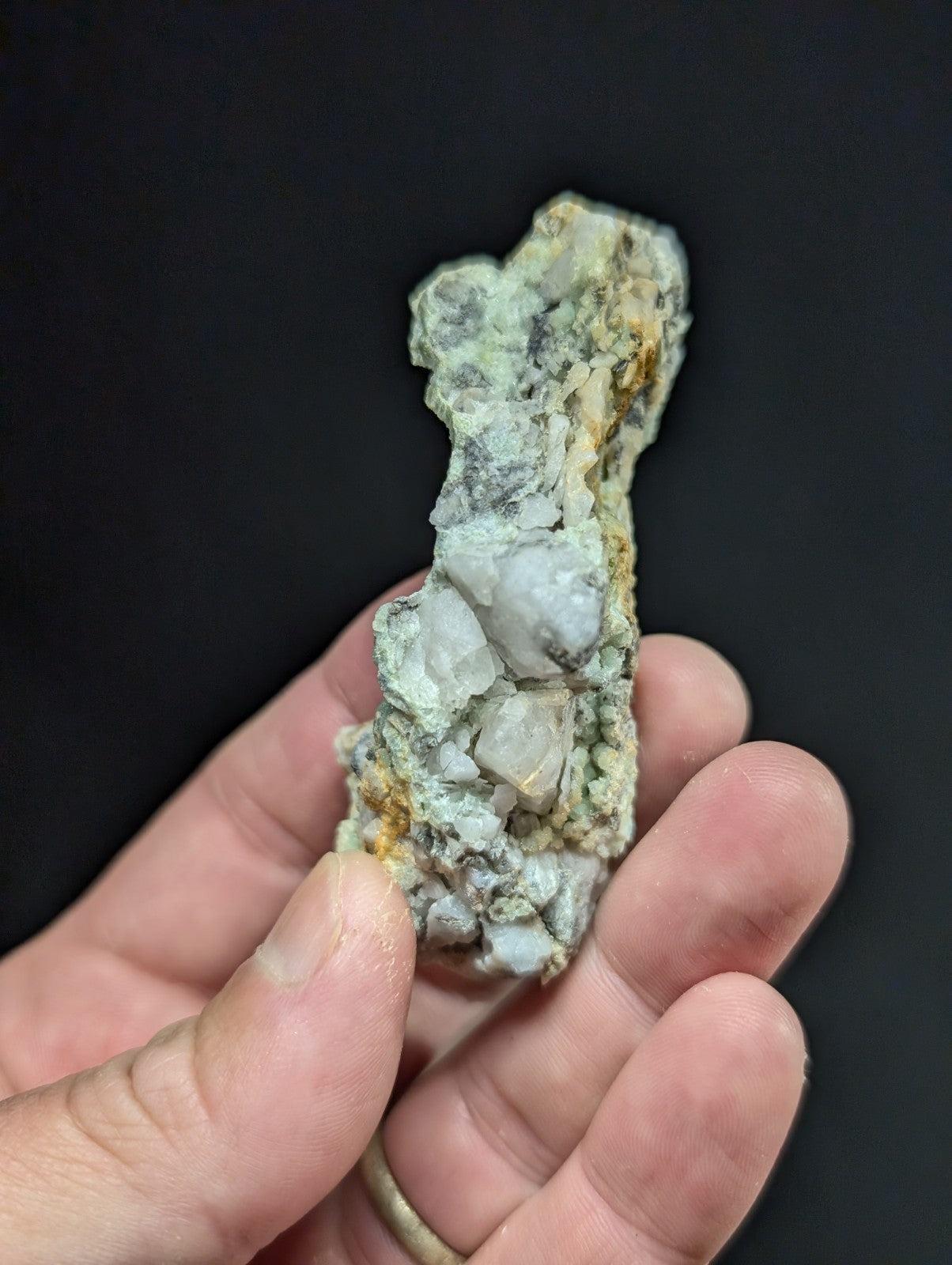 A hand holds a rough, irregularly shaped mineral specimen featuring translucent white crystals embedded in a matrix of gray and yellowish rock.