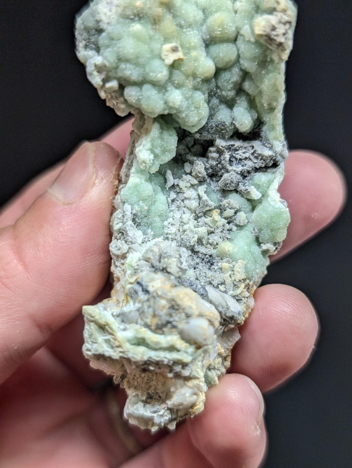 A hand holds a rough, irregularly shaped mineral specimen featuring clusters of pale green crystals intermingled with gray and yellowish matrix material.