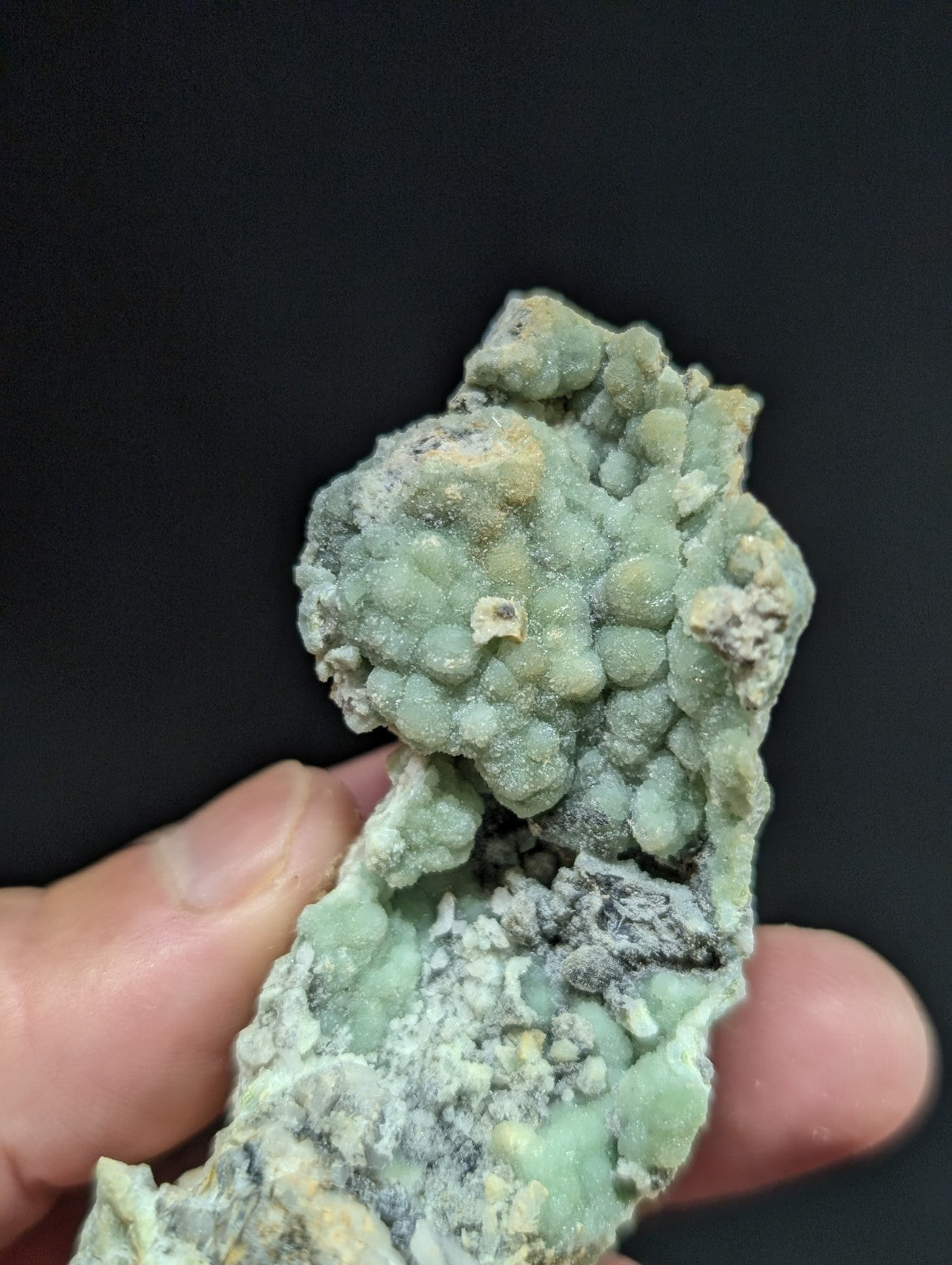 A textured, light green mineral specimen with a bumpy, clustered surface and patches of grayish material.