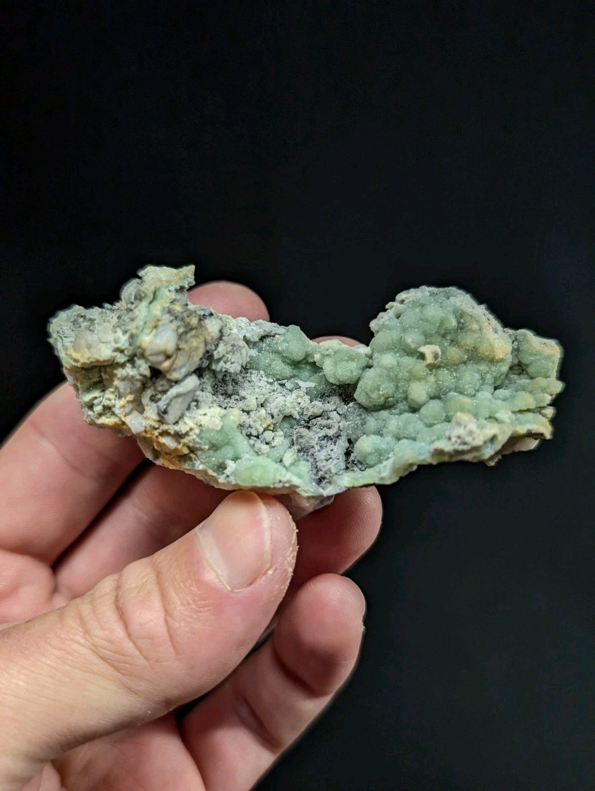 A hand holds a pale green, textured mineral specimen with patches of gray and yellowish hues.