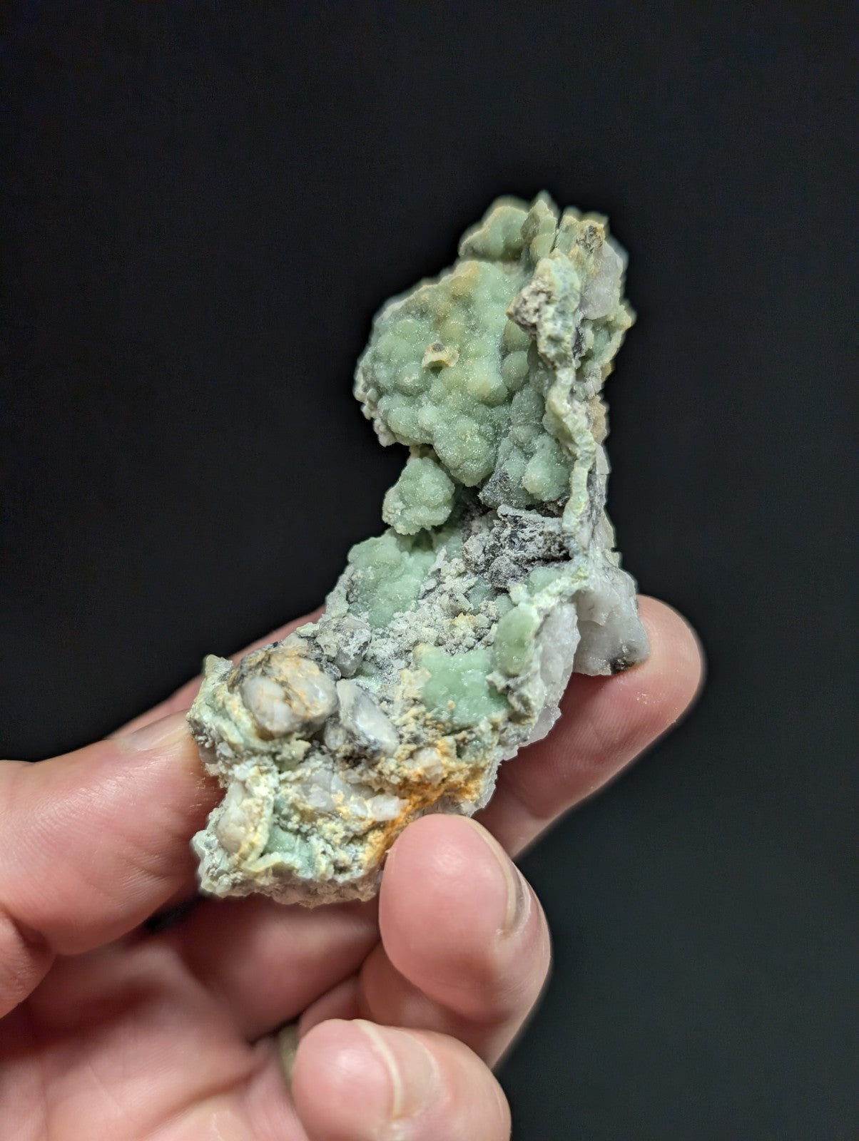A hand holds a rough, irregularly shaped mineral specimen with patches of pale green and grayish-white material.