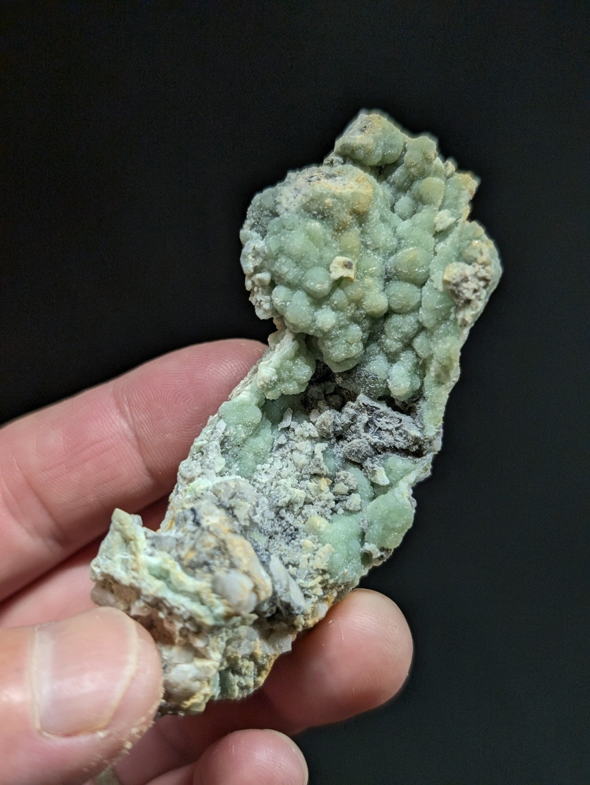 A hand holds a light green, bumpy mineral specimen with patches of gray and yellowish material.