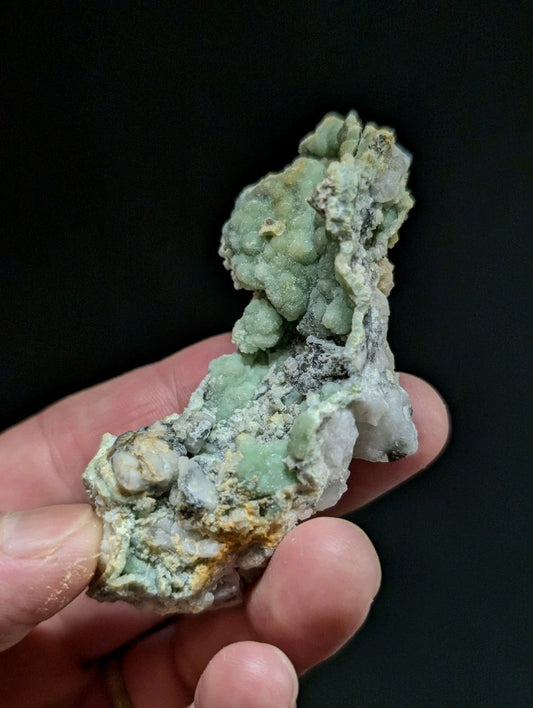 A textured, light green mineral specimen with patches of white and yellowish hues held in a hand.