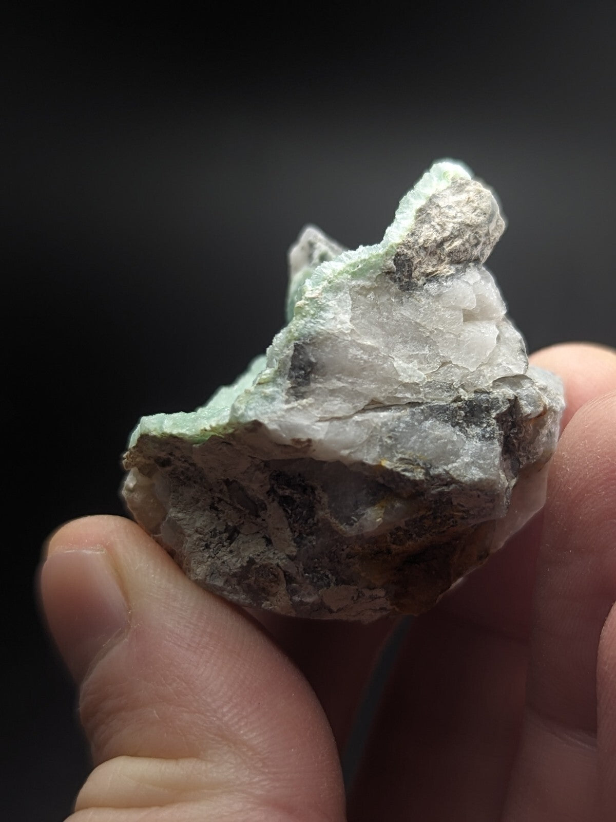 A hand holds a rough, irregularly shaped mineral specimen with translucent white crystals and patches of greenish-blue veining against a dark background.