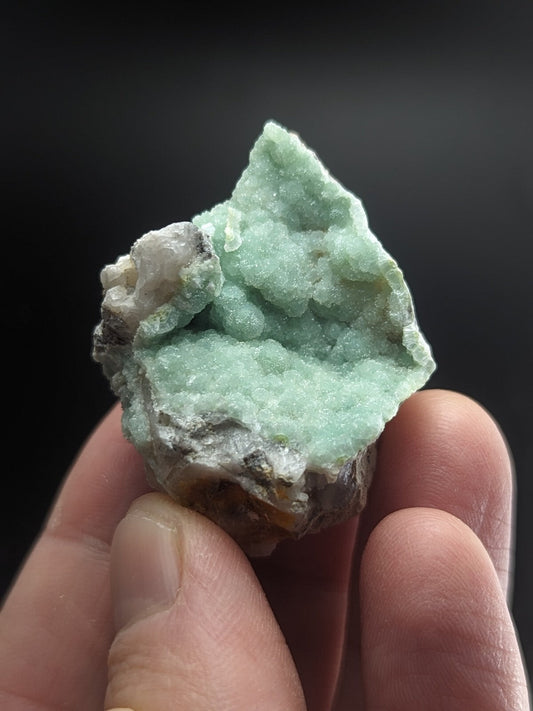 A hand holds a light green, crystalline mineral specimen with rough, jagged edges and patches of white and gray matrix.