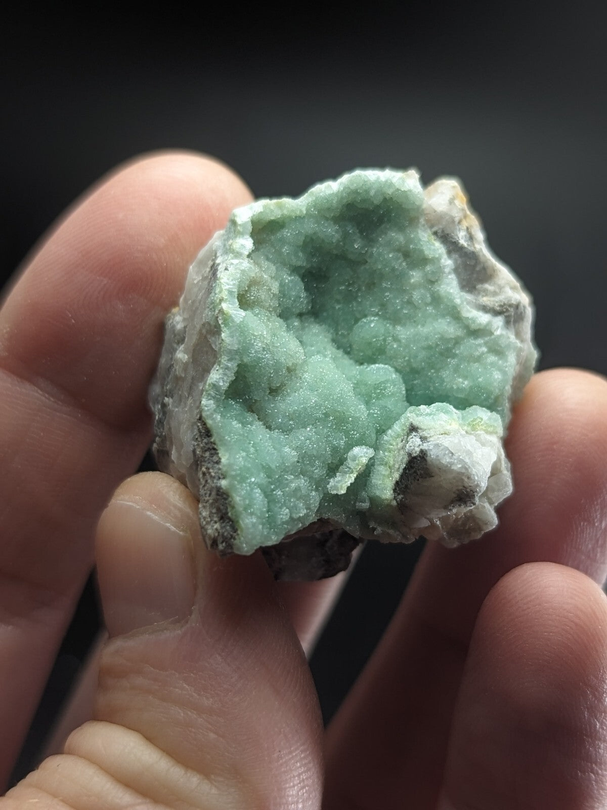 A hand holds a rough, light green mineral specimen with crystalline formations and white matrix.