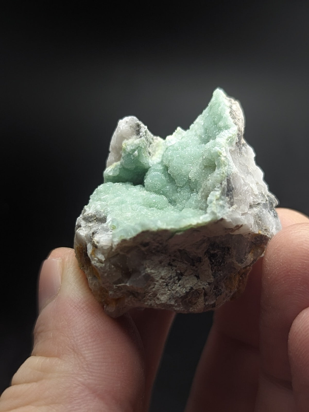 A hand holds a rough, irregularly shaped mineral specimen featuring vibrant light green crystalline formations emerging from a darker gray matrix.