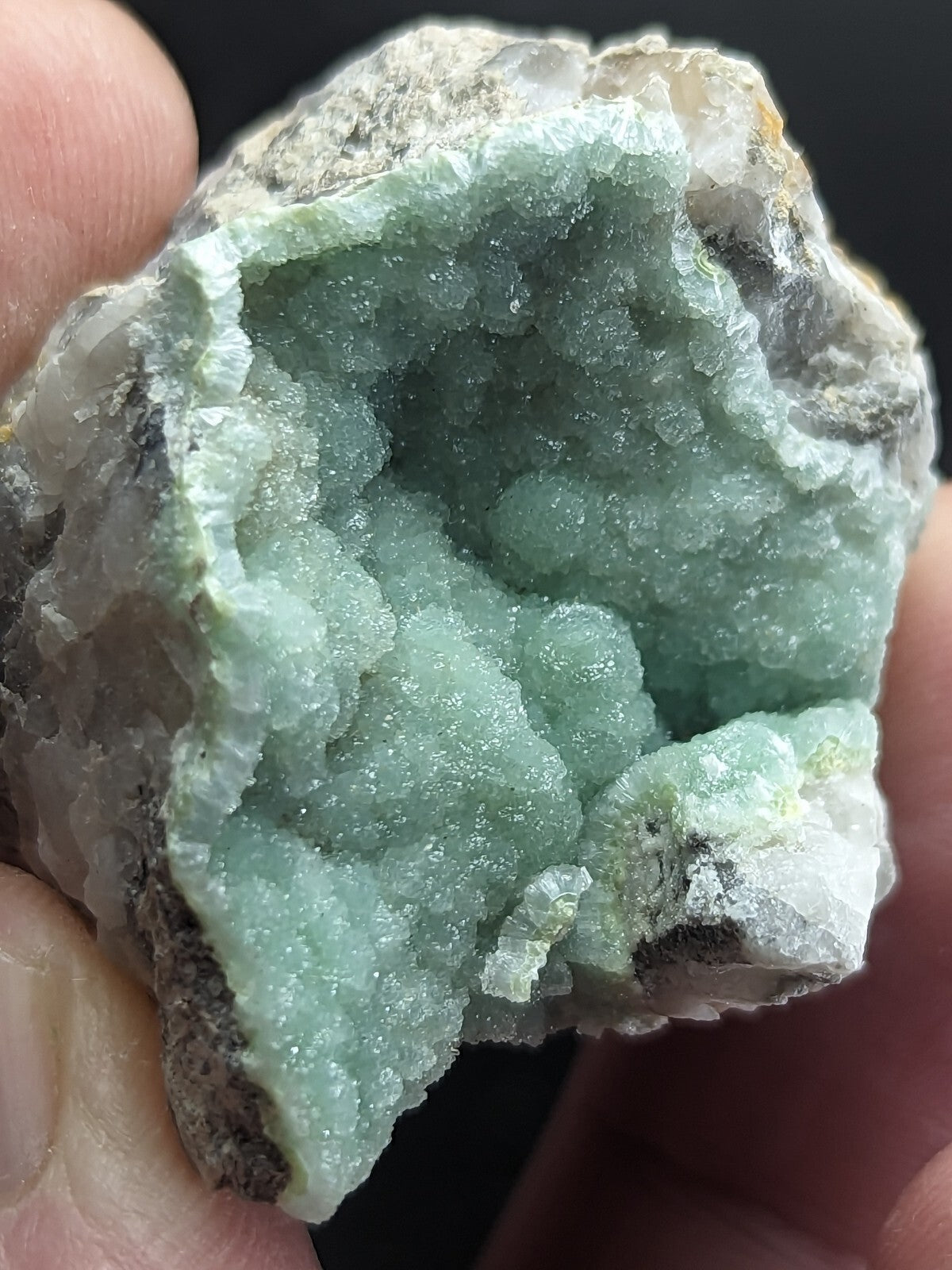 A hand holds a geode with translucent, light green crystalline formations inside and a rough, grayish outer shell.