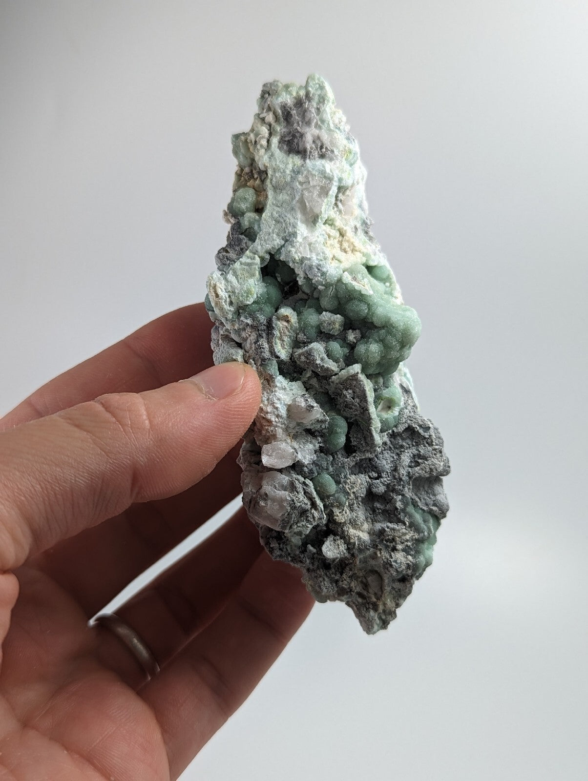 A hand holds a textured mineral specimen featuring clusters of greenish-blue crystals growing from a gray, rocky base.