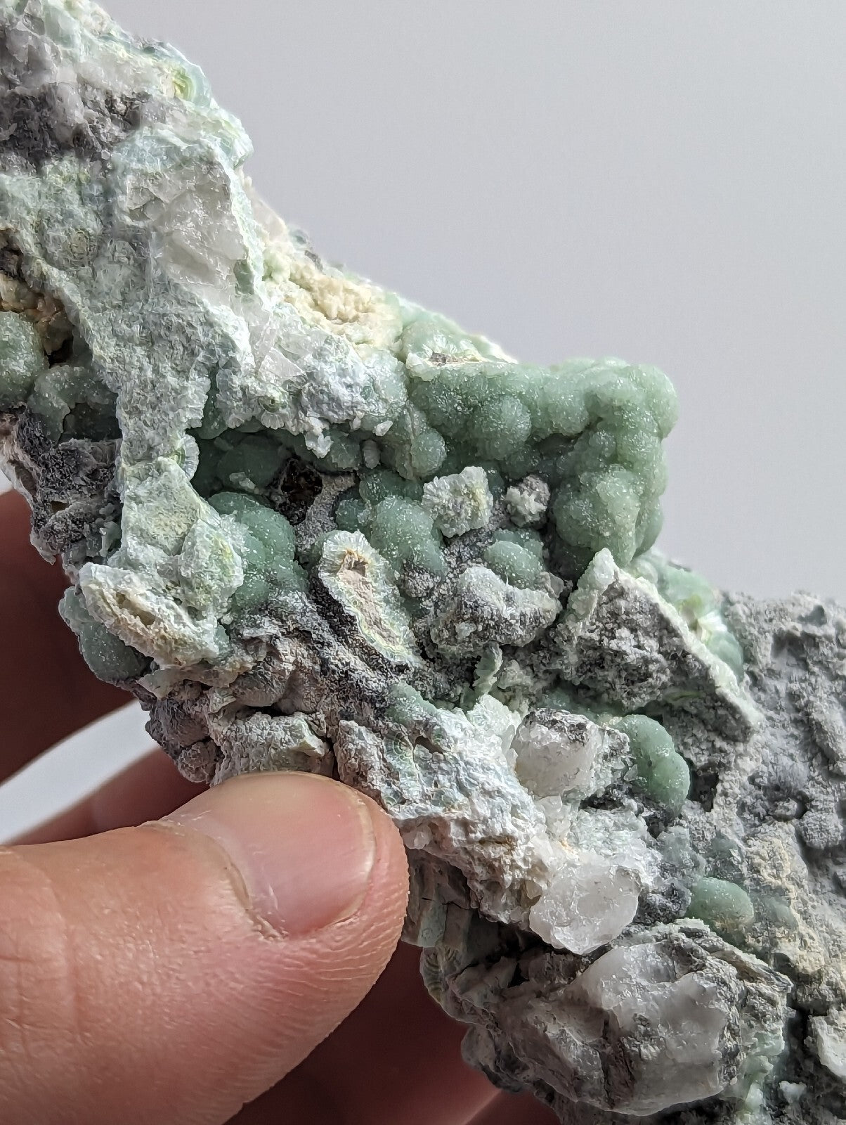 A hand holds a mineral specimen featuring clusters of light green, rounded crystals growing from a grayish matrix with white and translucent patches.